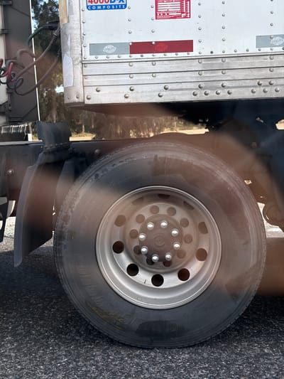 A close-up depicts a truck tire, mounted on a silver rim with circular holes. The tire is worn and branded "Bridgestone". The truck itself is partially visible, featuring a white trailer with vertical seams and rivets. A black mud flap is positioned behind the tire. The background shows a blurry mix of foliage and pavement, suggesting an outdoor setting.