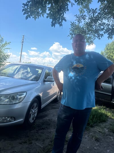 The image shows a man standing next to a silver car on a sunny day, with a bright blue sky and white clouds visible. He is wearing a light blue t-shirt with a graphic on the chest that reads "EXPLORER NATURE IS CALLING" in a circular design featuring a stylized wave and sun. The man is bald and wearing glasses. Trees are visible in the background, and the ground appears to be dirt and grass. A utility pole with power lines is also visible in the distance. The scene suggests an outdoor setting, possibly in a rural or suburban area, given the car, utility pole, and natural surroundings. The overall mood appears relaxed and casual.