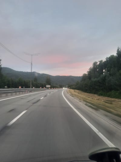 The image depicts a road in a rural, mountainous area, likely in Rybalka, Russia. The perspective is from a vehicle moving along the road, indicated by the blurred foreground and the slight tilt of the camera. The road itself is paved with a white line marking the lanes. To the right of the road, there is a grassy verge. The left side of the image shows a dense forest covering the slopes of hills or mountains. In the distance, the sky is a soft, overcast grey with subtle hints of pink and orange, suggesting either dawn or dusk. There are some lights visible along the roadside in the distance, and a solitary radio tower or antenna can be seen against the sky on the left. The overall atmosphere is calm and serene, with the overcast sky and muted colors contributing to a tranquil mood. There is no visible text in the image.