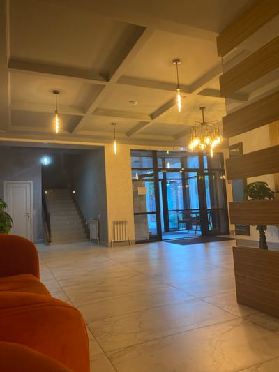 The image shows the interior of a modern building, likely a hotel lobby or common area, in Оқжетпес, Kazakhstan. In the foreground, a portion of an orange sofa is visible. The floor is tiled with a light-colored marble pattern.
The space features a coffered ceiling with recessed lighting and several decorative pendant lights. To the left, a staircase with grey steps and black railings ascends. Adjacent to the staircase is a white door.
The central area of the image shows a glass-enclosed entrance with double doors. Through the glass, two chairs and a table are visible, suggesting an outdoor seating area. The walls have a textured finish, and a modern radiator is mounted on the wall near the staircase.
To the right, a large wooden decorative element with horizontal slats is present, partially obscuring a potted plant. The overall ambiance is contemporary and well-lit, with warm-toned lighting from the fixtures. No individuals are visible in the scene.