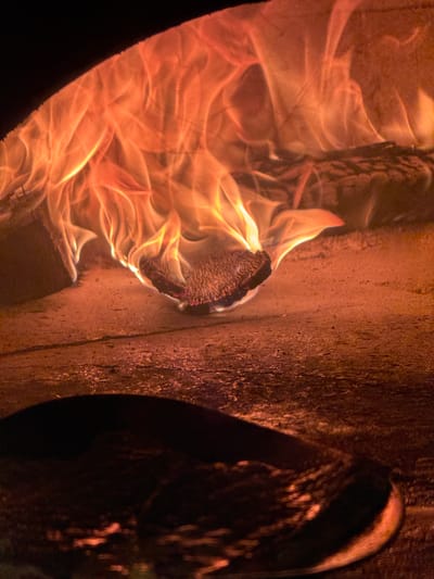 The image shows a close-up of a wood-fired oven with flames. The focus is on the glowing embers and the dancing flames, which are orange and yellow. The floor of the oven appears to be made of stone or concrete and is covered in a fine ash. In the foreground, out of focus, is a dark, circular object, possibly a pizza peel or a baking stone. The scene is dimly lit, with the fire being the primary source of light. The environment suggests a culinary setting, likely a restaurant or a home kitchen with a specialized oven. The image does not provide any information about the city of West Palm Beach, United States of America.