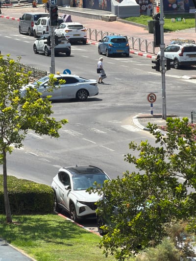 This is a high-angle, eye-level medium shot of a street intersection in Ashdod, Israel. The scene is captured on a sunny day. The street is paved and appears to be a multi-lane road with painted lane dividers and pedestrian crosswalks.
In the foreground, a white SUV with a black roof is parked partially on a patch of green grass, next to a neatly trimmed hedge. To the left of the SUV, a tree with vibrant green leaves casts shadows on the grass. In the middle ground, a white taxi with the markings "Gett" and "19317" is stopped at the intersection. Another white car is behind the taxi. Further back, a blue hatchback is in the right lane, and a white SUV is in the left lane.
A pedestrian, an older woman wearing a white shirt and dark shorts, is walking across the crosswalk at the intersection, carrying a shopping bag. Traffic lights are visible at the intersection. A "no U-turn" sign is also present. In the background, there are buildings and more vehicles, including a silver minivan and two white vans. The overall environment suggests an urban setting.