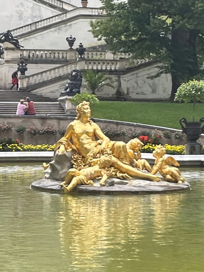 The image captures a golden sculpture of a reclining male figure surrounded by cherubs, situated in a pond. In the background, a grand, multi-tiered staircase leads up to a building. The staircase is adorned with ornate railings and several black sculptures, including a lion and various urns. Two individuals are seated on the staircase, facing away from the camera. A woman in a red skirt and black top is descending the stairs. Green grass and trees surround the area, with yellow and red flowers planted along the lower edge of the staircase. The overall scene suggests a public garden or palace grounds. This sculpture is located in the gardens of Ettal Abbey in Ettal, Germany.