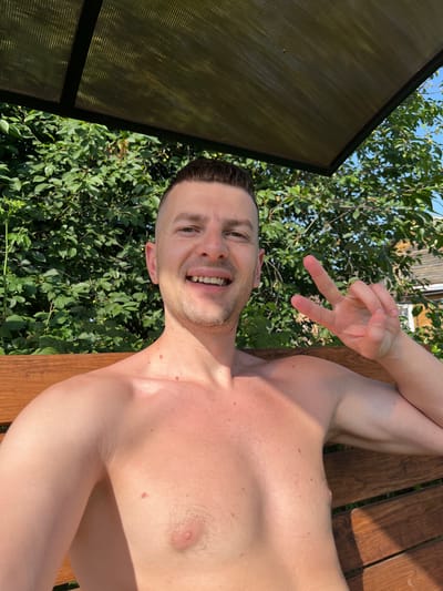 A man with short brown hair smiles at the camera, making a peace sign with his right hand. He is shirtless and appears to be sitting on a wooden bench in an outdoor setting. Behind him is a dense green hedge, and above him, a transparent roof supported by a dark frame. Sunlight casts shadows on his chest and arm. The location is identified as Navahrudak, Belarus.