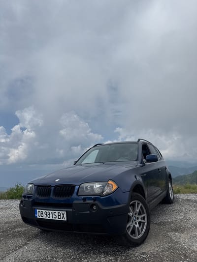 A blue BMW X3 SUV is parked on a gravel surface with a hilly, mountainous landscape in the background under a cloudy sky. The car's license plate reads "OB 9815 BX". The scene appears to be a scenic overlook, possibly in the city of Hristo Danovo, Bulgaria, given the license plate country code. There are no people visible in the image.