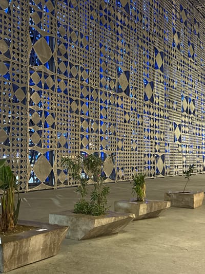 The image shows a modern building facade in Kruja, Albania, illuminated by blue lights at night. The facade features a decorative metal screen with a geometric pattern of diamonds and triangles, creating a lattice effect. Behind the screen, the blue lighting highlights the architectural structure and suggests windows or openings. In the foreground, several concrete planters hold small trees and plants. The ground is paved with concrete. The scene is devoid of people.