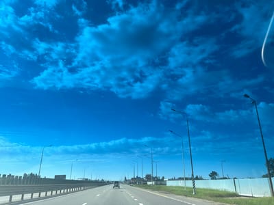 The image depicts a highway in Tlustenkhabl, Russia, under a bright blue sky with scattered clouds. A light-colored sedan drives in the distance in the center lane. Roadside streetlights line both sides of the highway. A barrier runs along the left side of the road. A fence and vegetation are visible on the right side. The road surface has lane markings and appears to be asphalt.