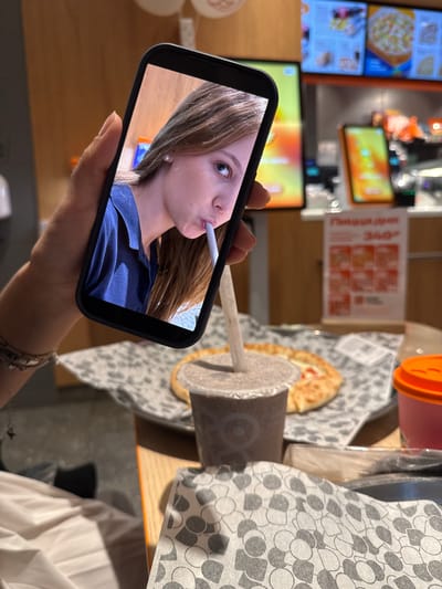 A person is holding a smartphone in their left hand, displaying a selfie of a young woman with light brown hair, wearing a navy blue shirt and pearl earrings, drinking from a straw. The smartphone is positioned in the foreground, partially obscuring a table with food and drinks. On the table, there is a half-eaten pizza on a patterned paper wrapper, a clear plastic cup with a brown beverage and a straw, and an orange-lidded cup. In the background, out of focus, are digital menu boards and restaurant interior elements. The overall scene suggests a casual dining setting, possibly a fast-food restaurant. The image quality is somewhat grainy. There is no discernible information in the image that would indicate the location is Chusovoy, Russia.