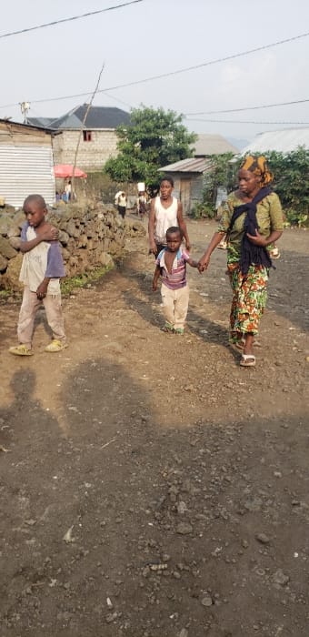 This image depicts a daytime scene on an unpaved dirt path in Buzove, Democratic Republic of the Congo, against a backdrop of modest residential structures and sparse vegetation under a light sky.

In the immediate foreground, the path is composed of packed dirt and small stones. To the left, a low wall constructed from dark, irregularly shaped rocks is present.

Four primary individuals are visible in the mid-ground. On the right, an adult female with a patterned headwrap, a green patterned top, and a floral long skirt, wearing white sandals, walks while holding the right hand of a young child. A baby, partially obscured by a dark fabric carrier, is secured on her back. The young child, wearing a striped shirt (pink, purple, white) and light-colored pants, with light green footwear, is walking beside her. Directly behind this pair is an older child or young adult female in a white sleeveless top and dark lower garments. To the far left, an older child or young male, dressed in a light-colored long-sleeved shirt (partially blue) and light-colored pants, with yellow sandals, stands near the rock wall, facing left but looking towards the right.

In the background, multiple structures are visible, including buildings with corrugated metal roofing and walls, and more substantial cinder block or stone buildings, one featuring a dark roof and a visible red-framed window. Overhead power lines are discernible against the sky. Further down the path, several other individuals are present, some appearing to carry white containers, possibly water jerrycans. A red umbrella or awning is also visible in the distance. The overall environment suggests a residential area with unpaved roads and mixed construction styles.