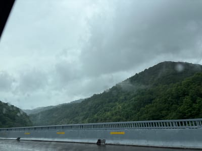 An exterior, eye-level, long shot captures a highway view in the region of Achara, Georgia. The image is dominated by a gray, overcast sky with visible clouds. In the foreground, a wet highway barrier with reflective yellow markings lines the right side of the frame. Beyond the barrier, lush green, tree-covered mountains rise, partially shrouded in mist in the distance. The wet road surface suggests recent rainfall. No people or specific vehicles are discernible in the image.