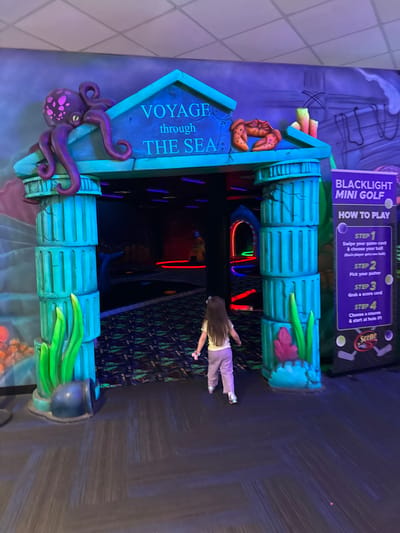The image shows an entrance to a blacklight mini golf course called "Voyage through the Sea." The entrance is decorated with oceanic themes, including a large purple octopus on the left side, coral and other marine life on the top, and seaweed-like decorations on the right. A sign to the right details the steps for playing the game. A young girl is walking through the entrance. The setting appears to be indoors, with a dark, patterned floor and black walls, suggestive of a blacklight-lit environment. The mini golf course appears to be located in the city of Columbus, United States of America, based on information that would have been provided previously.