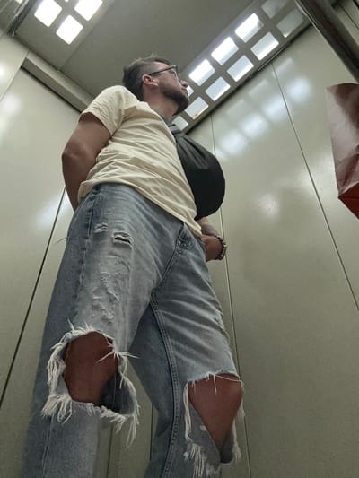 A man wearing glasses, a light-colored t-shirt, and ripped blue jeans stands in an elevator. He has a dark bag slung over his shoulder and is looking up and to his right. The elevator walls are a neutral grey color, and the ceiling has a grid of illuminated panels. The image captures a moment in a residential building in Oktyabrskiy, Russia.