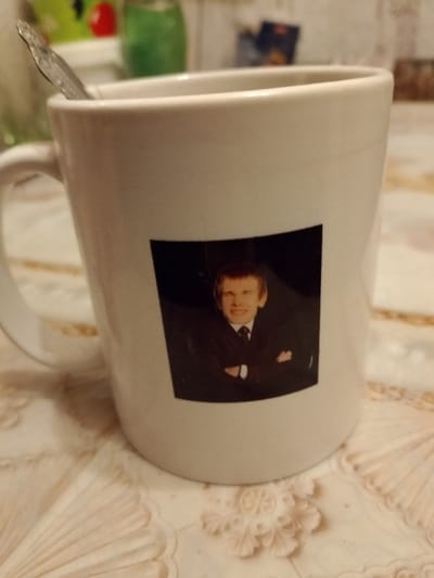 A white mug sits on a beige patterned surface. A silver spoon is visible inside the mug. A black square sticker adorns the mug, displaying a portrait of a man in a suit with his arms crossed. The background shows a blurry kitchen setting with green and red items.