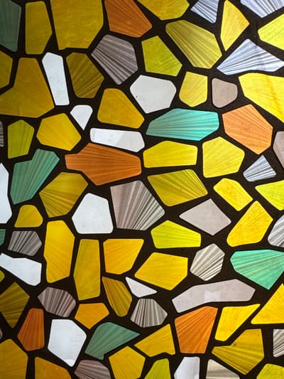 The image shows a close-up view of what appears to be a decorative window or panel. The design features irregularly shaped pieces of colored glass or translucent material, resembling a mosaic or stained glass pattern. The pieces are primarily yellow, orange, blue-green, white, and gray, separated by a dark brown or black grid. The pieces feature textures that look like converging stripes, and have varying levels of opacity. The image does not contain any people or other objects other than the panel. The location of the image is not possible to determine from the visual elements.