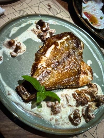 A close-up, overhead shot displays a beautifully plated dish of fish served with mushrooms and a creamy sauce. The fish, likely a sole or flounder due to its flat shape, is pan-seared to a golden-brown crisp, with visible scales creating a textured, appealing surface. It is positioned prominently in the center of a round, sage-green plate.
Surrounding the fish are several pieces of cooked mushrooms, nestled in a rich, pale sauce that appears to be a cream-based sauce, possibly infused with herbs or garlic, as evidenced by small flecks within it. A few sprigs of fresh basil, a vibrant green contrast, are placed artfully over the fish and sauce, adding a touch of freshness and aroma to the presentation. The plate itself rests on a surface that appears to be a table covered with a patterned paper placemat. The placemat has abstract line designs in a muted beige and brown color palette.
In the upper left corner of the image, a glimpse of another dish is visible in a white, shell-shaped serving dish. This dish contains what looks like thinly sliced raw fish, possibly sashimi, garnished with small green specks and a reddish element, possibly roe or a herb.
The lighting suggests an indoor setting, likely a restaurant, with warm ambient light contributing to the rich colors of the food. There are no people visible in the frame. The overall mood is one of fine dining, with attention to detail in the presentation of the food. The visible text on the placemat is partially obscured, but includes numbers like "010" and fragments of what appear to be foreign words, such as "fici". The location context provided is Denver, United States.