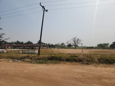 The image displays an expansive outdoor scene, likely a community field or sports ground, under bright daytime sunlight in Umuoye, Nigeria.
In the immediate foreground, a reddish-brown dirt road or path stretches across the width of the frame, showing signs of wear and loose soil. The ground transitions into a dry, grassy area with scattered debris and sparse vegetation.
A prominent feature in the mid-ground is a tall, dark wooden utility pole positioned centrally, with several overhead power lines extending across the upper part of the sky. To the right of the pole, a metal goalpost frame, characteristic of a football pitch, stands on the open, dusty field. The field itself is largely brown and dry, with patches of withered grass, indicative of a dry season. Along the edge of the field, several white rectangular signs are visible, one of which clearly displays the text "TEACH THE MEDIA".
In the background to the left, a row of low-rise buildings with tiled roofs can be seen. These structures appear somewhat weathered, set against a backdrop of dense green trees, including palm trees. Further back and to the right, a large, deciduous tree stands out with its bare branches, further suggesting a dry period. The sky is clear and bright blue, with a noticeable sun flare in the upper right quadrant of the image.
No people are visible in the scene, which conveys a sense of quietude and possibly a temporarily unused or dormant sports facility within a semi-urban or rural landscape.