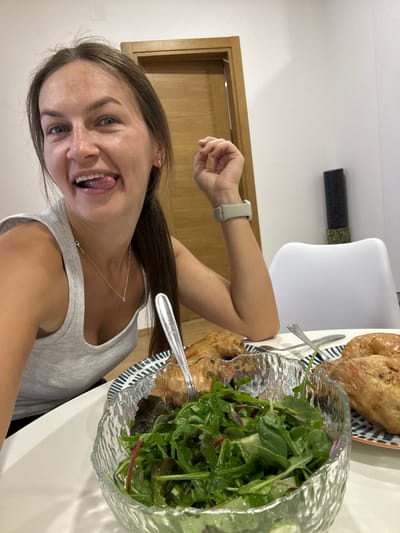 This is a vertical, eye-level, medium shot selfie of a young woman in Lapčići, Montenegro. The woman, with light brown hair and blue eyes, is sticking out her tongue and smiling widely at the camera. She is wearing a light gray tank top and a silver necklace. Her left arm is raised, and she's wearing a light gray smartwatch on her left wrist.

On a white table in front of her, there is a glass bowl filled with a green salad. To the right of the salad, there is a plate with two pieces of roasted chicken. A fork and knife are resting on the plate. The background shows a white wall and a wooden door. The lighting suggests it is daytime. The woman's expression and the food indicate a casual meal.