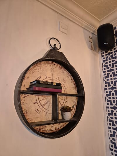 The image shows an overhead view of a decorative compass-shaped tray hanging on a wall, likely indoors in a residence in Breckland District, United Kingdom. The tray has a black, weathered-look frame and is divided into sections. Inside the tray, there are three small books leaning against a divider, and a small potted succulent plant in another section. The tray itself has a compass design printed on its surface, with markings like "NE," "MS," and degree measurements. Above the tray, on the upper left of the image, is a black "biamp." speaker mounted on the wall. The wall has a white or off-white paint, and there is a blue and white patterned decorative border at the top. A light switch is visible on the left wall, beside the compass.