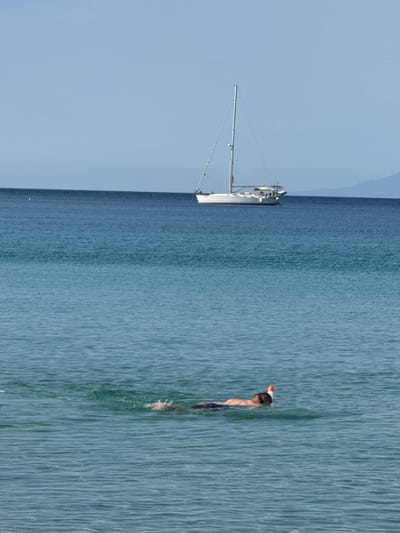 The image captures a serene scene on the water. In the background, a sailboat with its mast prominently displayed floats on the calm blue sea. The sky is a clear, pale blue, suggesting a pleasant day, likely midday given the lighting.
In the foreground, a person is snorkeling. They are partially submerged in the water, with their head, wearing a mask and snorkel, visible above the surface. The water is a deep blue, with ripples indicating gentle movement. The person appears to be enjoying the activity, perhaps exploring the underwater world. The overall impression is one of tranquility and leisure, a perfect summer day in a coastal location. The setting is a body of water, possibly the sea, with a hint of land or mountains in the far distance under the pale blue sky. No text is visible in the image.