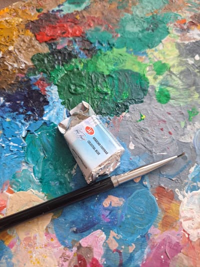 This is a close-up, overhead shot of an artist's palette laden with thick, colorful paint. The palette itself appears to be made of wood and shows signs of extensive use, with dried paint layered upon dried paint in a vibrant mosaic of blues, greens, yellows, reds, and purples. A silver-tipped paintbrush with a black handle rests diagonally across the palette, its bristles slightly splayed.

In the center of the image, a small, foil-wrapped watercolor paint pan is open, revealing a light blue pigment. The label on the paint pan is partially visible and reads "3K" within a red circle logo, followed by Russian text "НЕБЕСНО-ГОЛУБАЯ" and "CELESTIAL BLUE" in English. Below this, it indicates "ARTISTS WATERCOLOUR" and "3.5 ml". The paint pan is positioned as if recently used or about to be used.

The overall impression is one of a working art studio, possibly in Popovo, Bulgaria, where an artist is in the process of creating. The textures of the paint are prominent, suggesting an impasto technique or simply the build-up of layers over time. There are no people visible, but the presence of the tools and materials strongly implies an artistic activity. The lighting is bright and natural, suggesting daytime. The emotional tone is one of creativity and artistic exploration.
