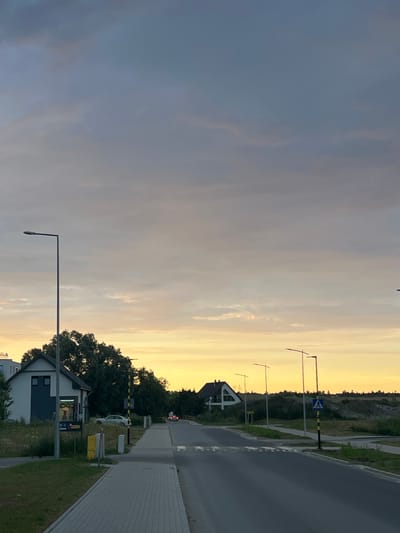 This media file shows a street scene in Rotmanka, Poland, at dusk. The sky is filled with soft, warm colors of yellow and orange from the setting sun, transitioning to a cooler gray on the left side of the image. Clouds are scattered across the sky, with some appearing more defined than others.
The scene depicts a residential street with houses visible on the left side. One house with a modern, angular design and dark-trimmed windows is prominent. A streetlamp with a minimalist design stands in the foreground, casting a faint glow. Further down the street, another house with a dark roof and windows is partially visible.
On the right side of the image, a paved sidewalk runs alongside the street. A road, likely a street or a small highway, is also visible. There are several streetlights with similar modern designs placed along the road. A pedestrian crossing is indicated by white markings on the road and a blue and white warning sign on a black and yellow striped pole. Trees and greenery line the sides of the road and surround the houses, suggesting a suburban or semi-rural setting.
A barrier gate, typically found at a railway crossing or a restricted area, is visible further down the road, along with a white car parked nearby. Yellow bins are also present. The overall atmosphere of the image is calm and peaceful, characteristic of the end of the day. There is no visible text in the image.
