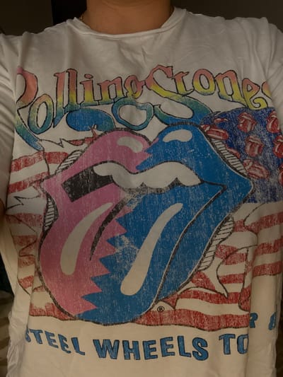 This is a close-up image of a white t-shirt with a graphic print. The graphic features the Rolling Stones logo, which includes a stylized pair of lips and tongue, along with the text "ROLLING STONES". There are also elements of the American flag incorporated into the design, with red and white stripes and a blue section with small tongue logos. Below the main graphic, the words "STEEL WHEELS TO" are printed in blue lettering. The t-shirt is being worn by a person, with a portion of their neck and collarbone visible at the top of the image. The lighting is dim, and the background is dark and out of focus. There is no discernible location in the city of Laval, Canada, visible in this image.