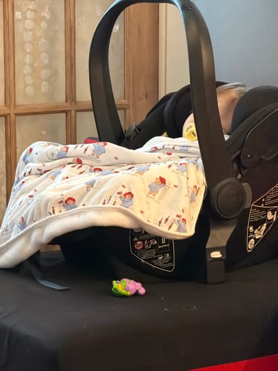 A baby, with a yellow pacifier in its mouth, is buckled into a car seat and covered with a Paddington Bear-themed blanket. A small toy is next to the car seat, against a black surface. The background shows a grid-patterned window. The car seat's information sticker is also visible.