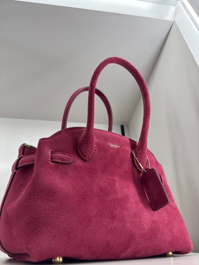 The image captures a close-up, low-angle shot of a maroon-colored suede handbag. The bag appears to be a Coach brand, with the logo subtly visible on the surface. The bag has two handles, and a small tag is attached. The bag is situated in a bright, presumably indoor, location, possibly a retail store, with white surfaces forming the background. The geographical location is identified as Sunrise, United States of America. No people or specific interactions are visible in the scene. The environment appears clean and well-lit.