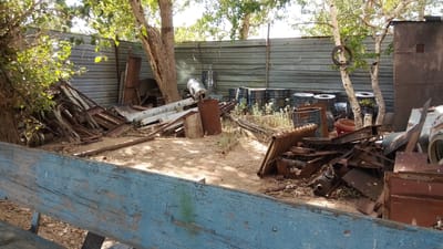 The image depicts an outdoor scene of a junkyard or scrap metal collection area, likely located in Kurchatov, Kazakhstan. The setting is characterized by a corrugated metal fence at the back, partially obscured by trees and foliage. Piles of rusty metal, including pipes, beams, and car parts, are scattered throughout the foreground and midground. Several black metal barrels, some appearing to be fuel or chemical drums, are stacked or lined up against the fence. A large, weathered blue wooden bench is prominently featured in the foreground, suggesting a viewing area or seating within the junkyard. The ground is covered with dirt and small stones. The lighting indicates it is daytime, with sunlight filtering through the leaves of the trees, creating dappled shadows on the ground. There are no people visible in the image. The overall impression is one of disarray and industrial remnants. No visible text is discernible in the image.