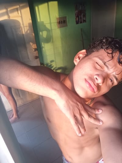 The media file is a selfie taken by a young man indoors, likely in La Guardia, Venezuela. The subject is shirtless, with curly dark hair and is looking towards the camera, winking. His arm is resting on his chest, and his hand is near his shoulder. The lighting suggests it is late afternoon or early evening, with warm sunlight streaming in from the left, casting shadows on a white door and a green wall. On the green wall, there are a few framed pictures. The floor appears to be tiled. In the background, to the left, another person's legs are visible, suggesting they are also in the room. The overall atmosphere seems casual and relaxed. There is no visible text.