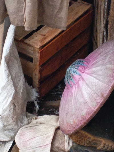 This close-up image presents a still life composition of various objects, suggesting a cluttered or storage-like environment in Welaluhu, East Timor.

The main subjects include a prominent wooden crate with visible grain and slat construction, positioned centrally and leaning slightly to the right. Its warm, reddish-brown tones contrast with the lighter elements. To the left, a large piece of white, crumpled fabric or a sack hangs downwards, displaying frayed edges near its base. Below it, more light-colored, folded or bunched textiles are visible, some appearing a pale pinkish-beige.

Dominating the right side of the frame is a vibrant pink, pleated object, likely a large, open fan, a parasol canopy, or a decorative hat. Its center features an intricate pattern of light blue and white shapes, adding a distinct ornamental touch. The pleats radiate outwards, giving it a soft, voluminous appearance. In the upper part of the image, a beige or light brown coarse fabric hangs, partially obscuring the top of the wooden crate. To the far right, a rough, textured, brownish material, possibly another fabric or a woven mat, is partially visible.

No activity or event is taking place; the scene is static. The lighting suggests an indoor setting or a deeply shaded area, with no direct sunlight. The textures of the wood, the varied fabrics—from rough sackcloth to the delicate pleats of the pink object—are notable details. There is no visible text in the image. The overall impression is one of everyday items, perhaps stored or casually arranged in a domestic or workshop space.