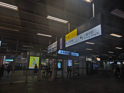 The image shows an interior view of a modern, well-lit transportation hub, likely a train station or subway station, in Kaohsiung, Taiwan. The architecture features a high ceiling with a suspended grid-like structure and numerous fluorescent light fixtures providing bright illumination. Large glass panels and doors dominate the scene, reflecting the lighting and some of the people present.

Several directional signs are visible, some in Chinese and some with English translations. One prominent yellow sign points to "Exit Jiouru 2nd Rd" in both languages. Another sign, black with white text and arrows, indicates "Sanfong Jhong St." to the left. Other signs for "Houyi Shopping Area" and "TR West Tickets" are also discernible. Inside the building, advertisements are displayed on illuminated panels, one featuring a bottle of what appears to be a beverage.

People are seen moving through the space. In the foreground, to the left, a group of individuals are walking past glass-fronted entrances. To the right, several people are walking in the darker, more open area of the concourse, with some individuals wearing masks. The flooring appears to be a dark, smooth material. The overall impression is one of a busy, functional public space designed for efficient transit.