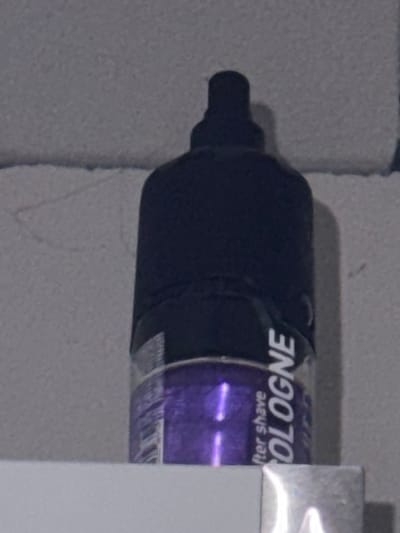 The image presents a close-up, horizontal view of a dark-colored bottle, likely an aftershave or cologne, resting against a light gray, textured surface. The bottle features a section with a purple hue, where text is visible. The text appears upside down and horizontally reversed, which, when corrected, reads "After Shave" and "COLOGNE". The left side of the bottle shows a dark, possibly black, cap or dispenser. A bright white, somewhat reflective object or surface partially obscures the right side of the bottle. The lighting in the scene is dim. There are no people visible, nor is any specific activity taking place. The setting appears to be indoors in Lelystad, Netherlands.