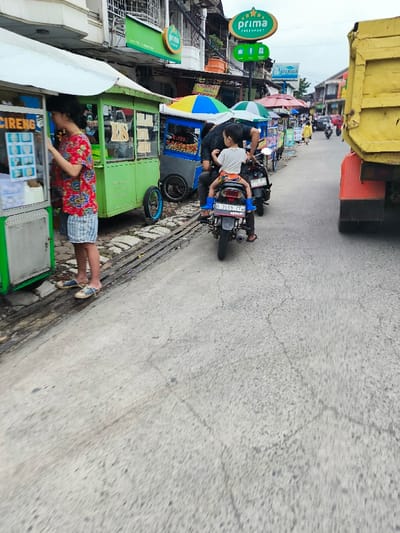 The scene takes place on a street in Kecamatan Dayeuhkolot, Indonesia, at the coordinates -6.963 latitude and 107.594 longitude. Three men and a young boy are visible in the image. The men are all wearing casual clothing. Two of the men are standing in front of food stalls, while the other man is on a motorcycle. The young boy is riding on the back of the motorcycle. The motorcycle is parked in front of a food stall. There are other food stalls and buildings visible in the background. There are some people walking in the background. The street is paved with concrete. The environment is a busy street scene.