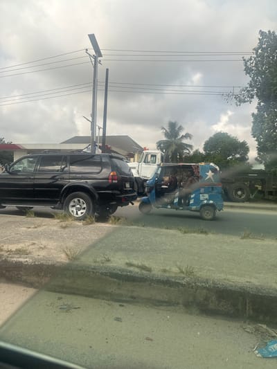 The image captures a street scene in Elimgbu, Nigeria, under an overcast sky. In the foreground, a black, four-door SUV, appearing to be a Mitsubishi Montero Sport, is stationary or parked, oriented to the left. Its rear bumper is visible, and it rests partially on a concrete median strip with sparse grass. To the right of the SUV, on an asphalt road, a blue and white three-wheeled auto-rickshaw, locally known as a "Keke Napep," is visible. This auto-rickshaw features a distinctive design with a white star on its rear and white stripes, and a male driver is visible within the cabin. Directly behind the SUV, a white heavy-duty truck is partially obscured. Further to the right, a dark green or black heavy-duty truck with a flatbed trailer is also partially visible on the road. Multiple tall utility poles with overhead power lines stand along the roadside; one pole is equipped with a solar panel and a light fixture. In the background, buildings with red roofs are visible, including one with a partially visible "MRS" sign, consistent with a gas station. Palm trees and other green foliage are present in the middle ground and background. The immediate foreground shows a dusty concrete surface and curb, viewed through a slightly tinted window.
