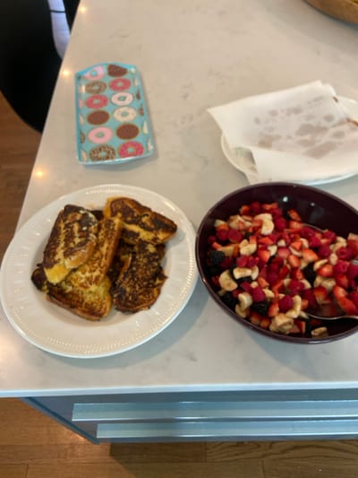 A white countertop in a kitchen in Rye, New York, located at coordinates (latitude: 40.952, longitude: -73.708), is shown. There are two white plates and a burgundy bowl on the countertop. One plate contains several slices of French toast, while the other has a piece of parchment paper. The bowl contains a fruit salad of strawberries, raspberries, blackberries, and bananas. A blue tray with a donut pattern is also present on the countertop. The countertop appears to be made of marble or quartz, with grey veins running through it. The countertop is situated above a grey cabinet with three drawers. The cabinet is situated above a wood floor, with wood paneling on the wall behind it. A black chair is visible in the background of the image. The countertop is clean and dry. There is no sign of use or activity in the image. There are no people or animals in the image. The image is well-lit and taken at eye level.