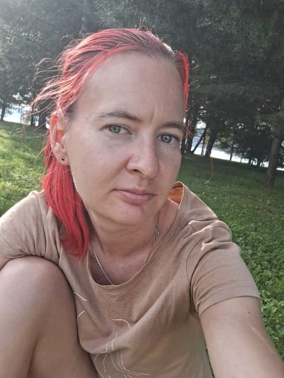 A medium shot taken outdoors shows a woman with bright red hair and fair skin. She is wearing a light brown t-shirt and a silver chain necklace. She has a small black stud earring in her left ear. The woman is sitting on green grass, with trees and a body of water visible in the background. The lighting suggests it is daytime, possibly late afternoon due to the long shadows cast by the trees. The overall impression is a candid portrait.