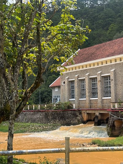 A vertical eye-level shot captures a building and a waterfall in Păng Tiêng 1, Vietnam. The building is constructed from grey stones and has multiple rectangular windows evenly spaced across its facade. It is topped with a red-tiled roof. Adjacent to the building, a waterfall cascades down a stone structure. The water is a murky, yellow-brown color. A rusty metal pole runs parallel to the waterfall. A tree with green leaves dominates the lower left corner of the frame, partially obscuring the scene. In the background, a dense forest can be seen. The lighting is bright and natural.