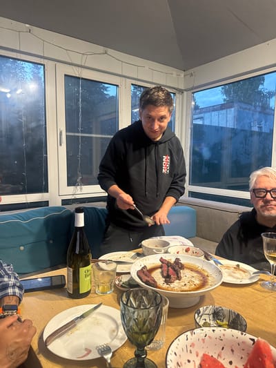 In Ptichnoye, Russia, a man in a black hoodie with "ZION" printed in red and white on the chest stands behind a wooden table, holding a spoon and a knife, appearing to serve food. He is looking towards the camera with a slight smile. To his right, another man with white hair and glasses, wearing a black hoodie, is seated at the table, also looking towards the camera. The table is set with plates of partially eaten food, including steak and watermelon, wine bottles, glasses, and cutlery. The setting appears to be an indoor dining area with large windows overlooking a twilight outdoor scene, possibly a sunroom or conservatory. Strings of fairy lights are visible across the top of the windows.