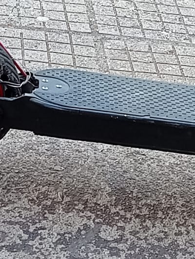 The image shows a close-up of an electric scooter, primarily focusing on the footboard. The footboard is black with a textured grey surface. The front of the scooter, including the tire and some wiring with red casing, are visible in the upper left. The scooter is positioned on a paved surface. The upper portion of the image features square paving stones, while the lower section displays a rougher, asphalt-like surface. The location is unspecified within Malko Chochoveni, Bulgaria.