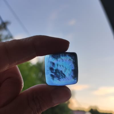 An extreme close-up, silhouetted shot shows a person's thumb and index finger holding a small, square, iridescent blue glass object. The object has a swirling, almost nebulous pattern within it and reflects light with a shimmering quality. The background is a soft blur of the sky and foliage, suggesting an outdoor setting. The sky is a gradient of pale blue, transitioning to a hazy yellow at the horizon, with wispy clouds. The foliage is a muted green. The overall lighting is soft, consistent with twilight or dawn. The image does not contain any discernible features that would allow for identification of the specific location within Kineshma, Russia.