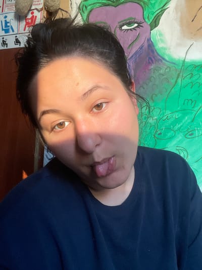 A young woman with dark hair pulled back in a messy bun is taking a selfie. She is wearing a dark blue crewneck sweatshirt. Her tongue is sticking out, and her eyes are looking at the camera. Sunlight is casting a shadow across her face, highlighting the right side of her face and her right eye. In the background, a colorful painting of a purple and green mythical creature is visible. To the left of the painting, there are several signs and objects on the wall, including what appear to be disability access symbols and some cylindrical objects. The scene appears to be indoors, possibly in a room in an apartment complex in ЖК «Борисоглебское», Russia.
