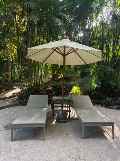 The image depicts an outdoor seating area situated within a lush, tropical environment, likely in Khlong Thom Nuea, Thailand. In the foreground, two light gray, modern-style chaise lounges are positioned on a light-colored, pebbled concrete patio. Between the lounges, a small, round table with a patterned cloth is visible, with a large, cream-colored umbrella propped open behind it, supported by a wooden pole. The umbrella's canopy casts shadows on the surrounding area. Behind the seating, a dense backdrop of various palm trees and other tropical foliage creates a dense, green canopy, with sunlight dappling through the leaves. The natural landscape features a winding path made of gravel and stones leading uphill to the left, with stone steps further up. The overall scene suggests a tranquil, resort-like setting designed for relaxation amidst nature.
