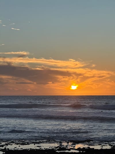 A serene coastal scene is captured, showcasing a radiant sunset partially obscured by wispy clouds in a pale sky. The sun's glow reflects faintly on the tranquil ocean waters, forming gentle waves. A dark, textured coastline occupies the right edge, suggesting rocks or a beach. The composition evokes a sense of calmness and natural beauty. The image is rotated 90 degrees.