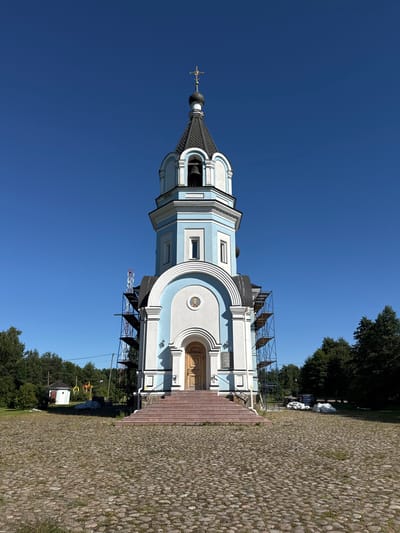 The image shows the Church of the Kazan Icon of the Mother of God in St. Ozerki, Russia, on a clear day. The church is a multi-tiered structure with a light blue and white facade, topped by a dark dome and a golden cross. Scaffolding is erected on the left side of the building, suggesting ongoing construction or renovation. The church is set on a cobblestone plaza, with a set of red-carpeted stairs leading to the entrance. To the left of the church, a small white building with a red roof is visible. The background consists of green trees and a clear blue sky. There are no people or animals visible in the image. Several white bags, possibly filled with materials, are piled on the ground to the right of the church.