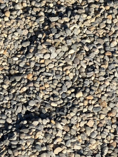 The image shows a close-up of a pile of small, rounded pebbles. The pebbles vary in color from light gray to tan and beige. The surface is densely packed with the pebbles. The location is unknown and Grad Opatija, Croatia has no relevance.