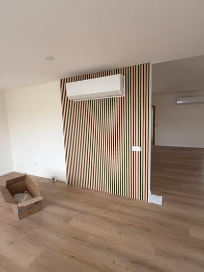 The image depicts an interior scene, likely a newly constructed or renovated residential space, in Gondomar, Portugal. The room features light-colored walls and a laminate wood-look floor.
Dominating the right side of the foreground is a decorative wall panel made of vertical wooden slats with dark vertical separations. Mounted on this panel, towards the top, is a white air conditioning unit. To the left of the panel, the wall is plain white and has a single electrical outlet.
In the left foreground, an open cardboard box lies on the floor, containing some crumpled packaging material. A small white cylindrical object, possibly a power adapter or a small appliance, is located near the wall to the left of the wooden panel.
The right side of the image opens into another room, also with a wooden floor and white walls. Another white air conditioning unit is visible on the wall in this adjacent room. A doorway with a wooden frame is partially visible on the left side of this adjacent room, leading further into the dwelling.
A piece of white paper or plastic sheeting is lying on the floor near the base of the wooden slat wall. The overall impression is of an unfinished or recently completed interior, with minimal furnishings. There are no people or animals visible in the image. The lighting suggests it is daytime, with ambient light entering from an unseen source.