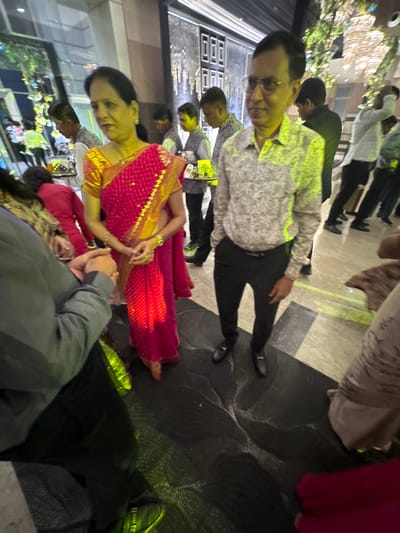 The image captures an evening social gathering or event, likely held in an indoor or semi-outdoor venue in New Town, India. The scene is bustling with people and illuminated by artificial lights, casting yellow and green light effects on the polished floor and subjects.

In the mid-ground, a man wearing glasses, a grey patterned shirt, and black trousers stands with a subtle smile, looking towards the left side of the frame. To his right and slightly closer to the foreground, a woman dressed in a striking red saree with intricate gold borders and details is visible. She has a warm smile and her hands are clasped together, suggesting a gesture of greeting or a traditional custom.

Numerous other individuals populate the background. Several men are seen in light-colored shirts or kurtas and dark pants, standing and engaging with one another. Some children are also discernible among the attendees. The background features decorative string lights, possibly adorning plants or architectural elements, and a reflective floor. In the immediate foreground, the shoulder and back of a person wearing a dark jacket or suit are visible on the bottom right.

The attire, particularly the traditional saree, suggests a formal or semi-formal occasion with a festive atmosphere. No visible text is clearly legible within the image.