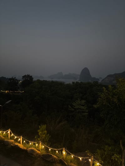 Captured at Khlong Khian, Thailand, the image presents a twilight scene. The left side of the frame is dominated by a cloudy sky. In the middle distance, silhouettes of hills rise above what appears to be a body of water. The right side of the image is dense with dark foliage and trees. String lights illuminate a path or ledge along the lower right edge, creating a visual contrast with the darkness.