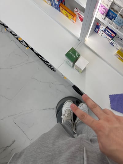The image shows a person's feet wearing white sneakers and gray sweatpants, standing on a marble-patterned floor. The person is holding up two fingers in a peace sign, with their hand and arm visible in the upper right portion of the frame. The background features shelves filled with various pharmaceutical products, including boxes and vials with labels in Cyrillic script. Some of the visible text includes "MUCOZYME," "Xyl," "АЦЦ," "Валиглог," and "Ксило." The products are arranged in a retail or pharmacy setting. The lighting appears to be artificial, suggesting an indoor location. The scene seems to be a casual moment captured during a shopping trip or visit to a pharmacy in Madan, Bulgaria.